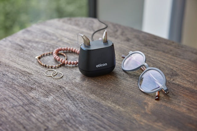 Zwei Oticon Intent Hinter-dem-Ohr-Hörgeräte in einer Ladestation neben Schmuck