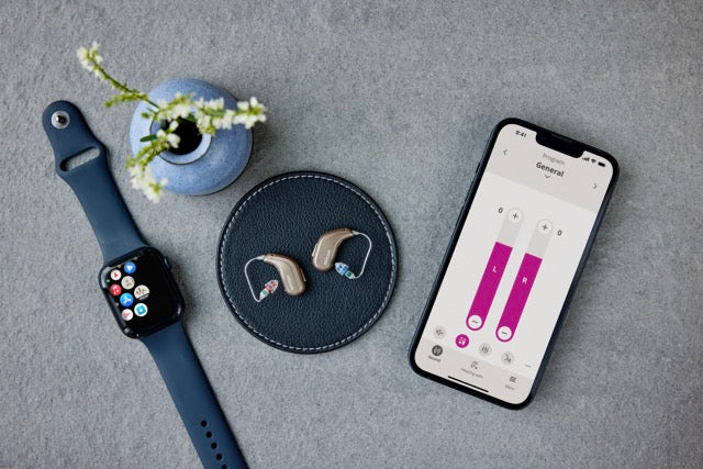 Zwei Oticon Intent Hinter-dem-Ohr-Hörgeräte neben einem Smartphone und einer Smartwatch